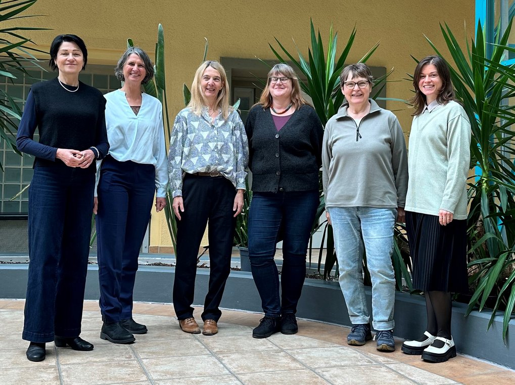 Sechs Frauen stehen nebeneinander im Innenraum, umgeben von Pflanzen.